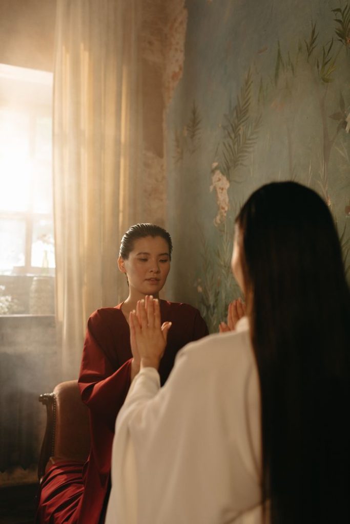 Two women meditating indoors in traditional attire with sunlight streaming through a window.